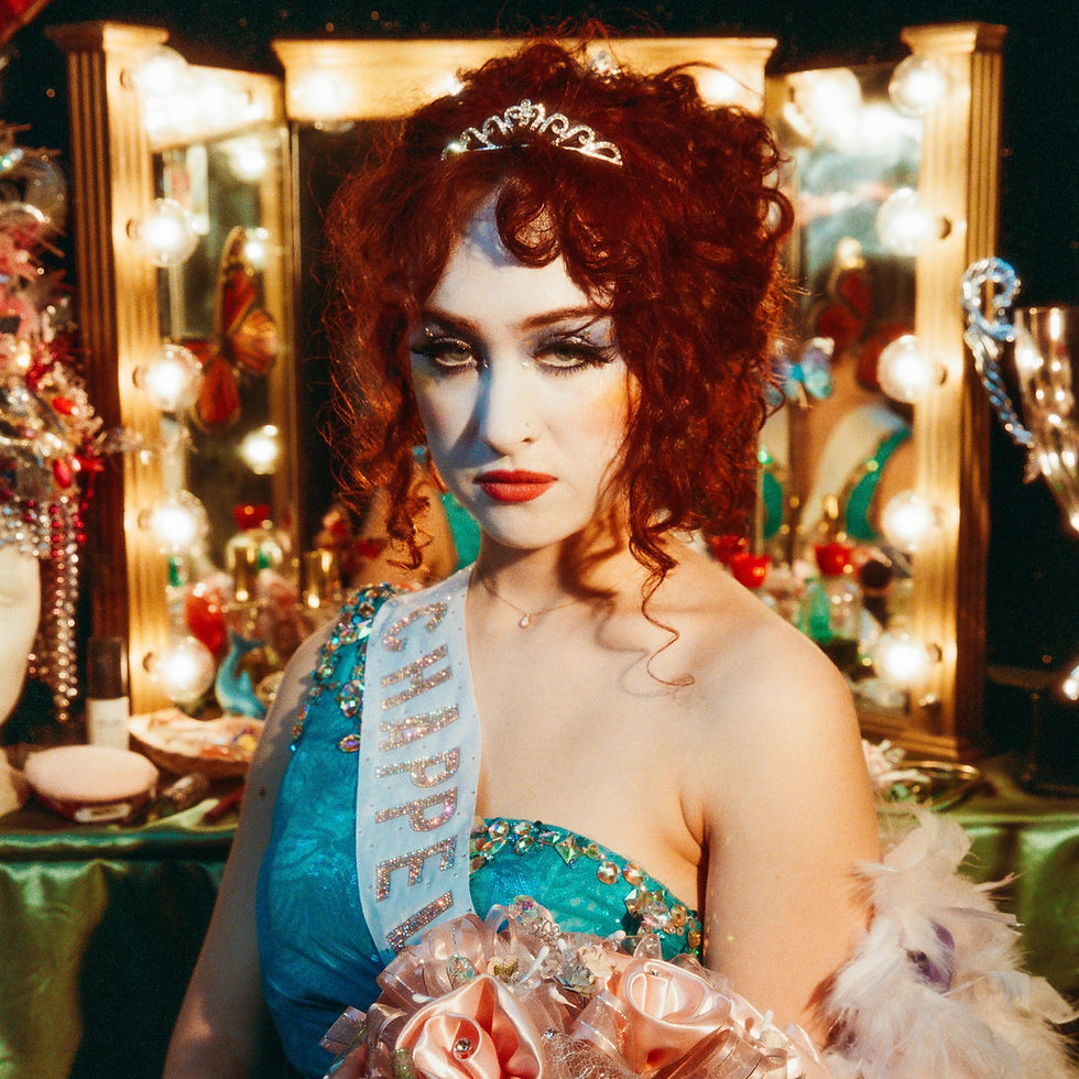 Red-haired Chappell Roan in blue dress with a tiara and a sash stands in front of a lit vanity mirror. Moody, vintage setting with floral bouquet.