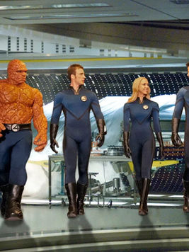 Four people (Fantastic Four: Rise of the Silver Surfer) in futuristic suits walk confidently in a high-tech, metallic room. One has an orange, rocky texture. Mood is determined.
