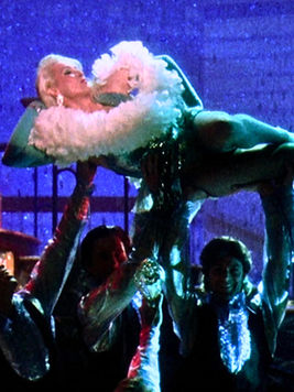 Meryl Streep (in Death Becomes Her (1992) ) in a feathered costume is lifted by dancers on a stage with sparkling blue lights, creating a glamorous, celebratory mood.