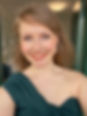 A portrait photo of Aimee who has dark blonde hair and blue eyes. Her hair is down and blowing backwards in the wind. She is smiling wearing lipstick and teal green asymmetrical dress off her shoulder. The background is blurry