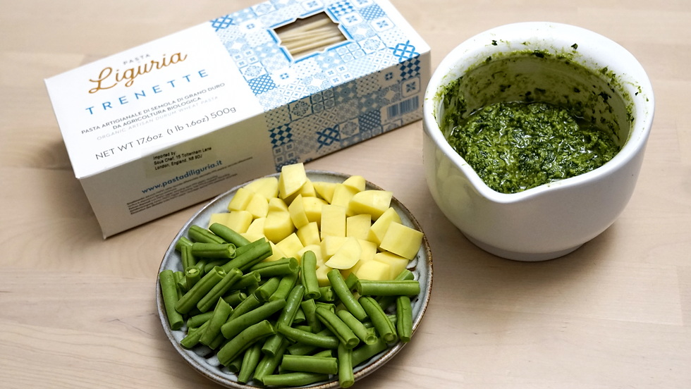 Box of Liguria trenette pasta, a bowl of green pesto, and a plate with chopped yellow potatoes and green beans on a light wooden table.