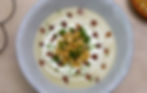 Creamy soup in a gray bowl topped with croutons, chopped chives, and bacon bits, with a swirl of cream. Wood table background.