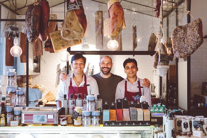 Three smiling people stand behind a deli counter with hanging meats above. They wear aprons in a warmly lit shop with various spices displayed.