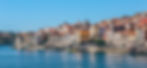 Colorful buildings with orange roofs line a waterfront in a sunny setting, with boats docked along the river. Clear blue sky above.