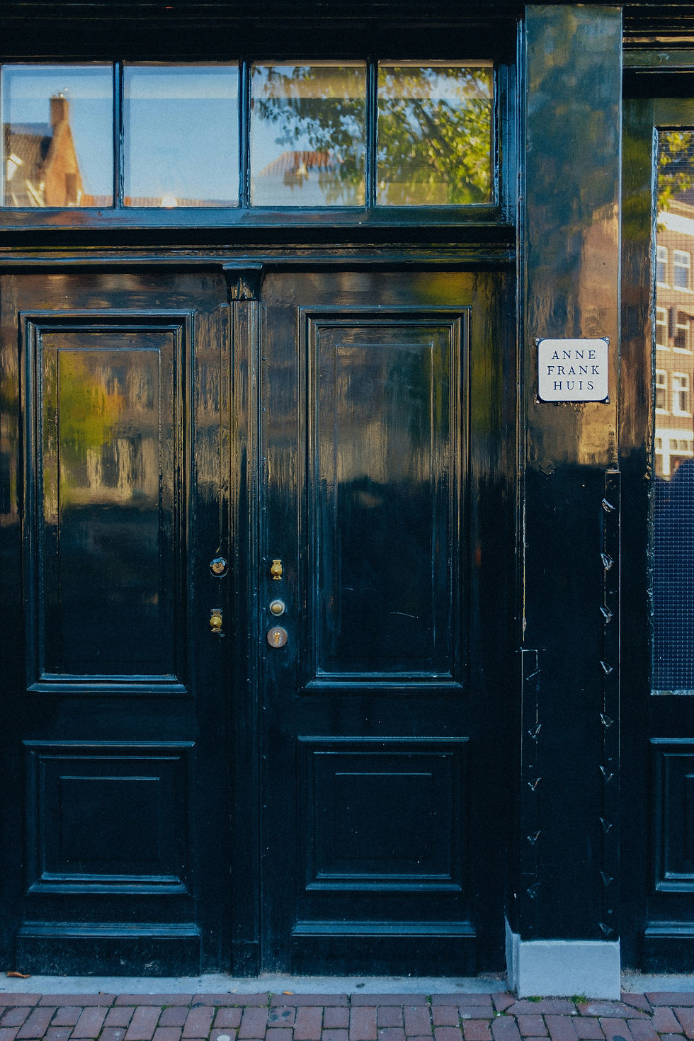Black double doors with windows at Anne Frank Huis, reflecting trees and buildings on a quiet street.