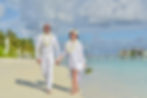 Couple in white attire holding hands, walking on a tropical beach with palm trees and blue ocean. Both wear flower leis, smiling.