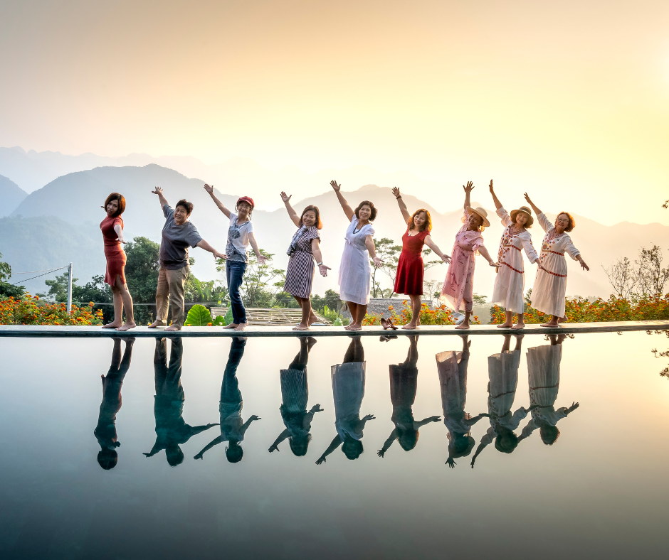 Nine people pose joyfully by a pool with vibrant outfits against a mountain backdrop at sunset, their reflections mirrored in the water.