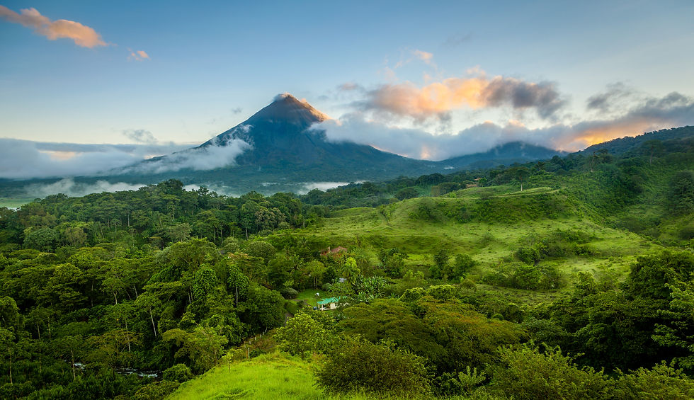 Lush green landscape with dense forest and rolling hills, dominated by a cloud-capped volcano under a clear blue sky with orange sunset hues.