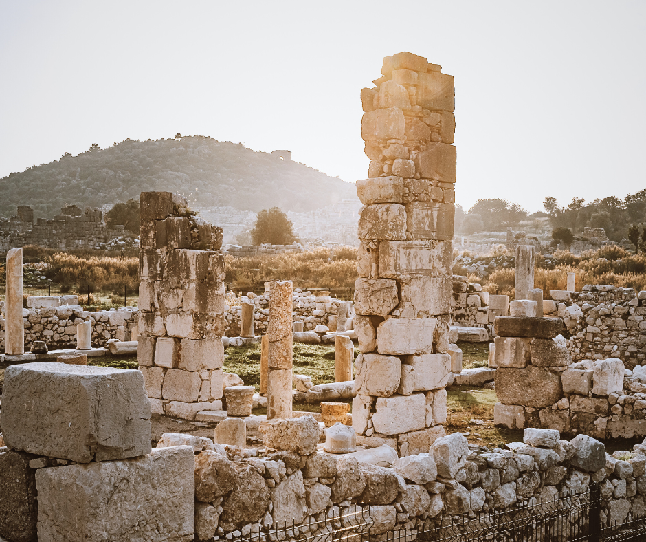 Ancient stone ruins with tall columns, set against a hilly landscape during sunset. Warm tones and scattered vegetation create a serene mood.