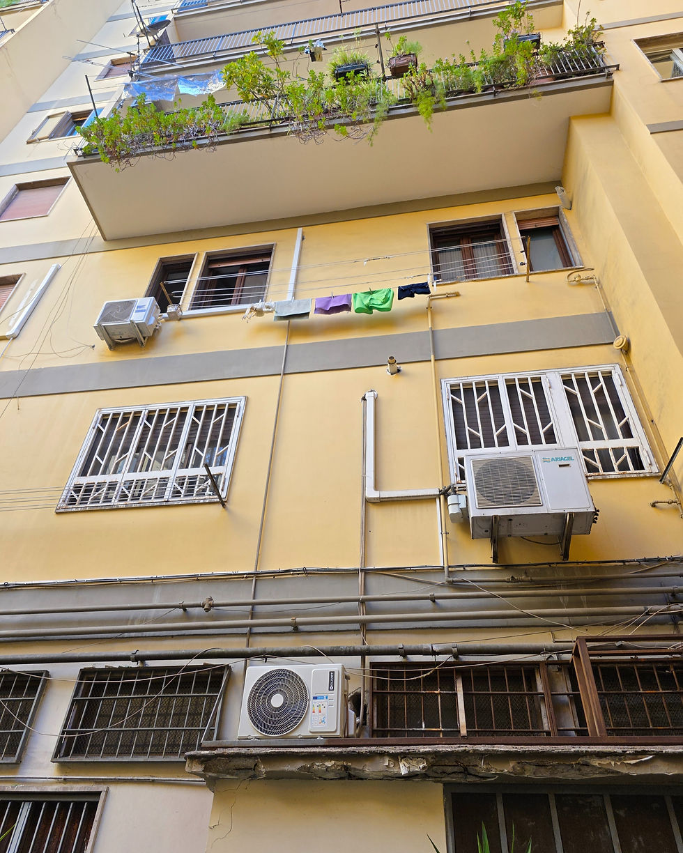 Yellow building facade with AC units, barred windows, and a balcony with plants. Clothes line holds colorful garments. Urban setting.