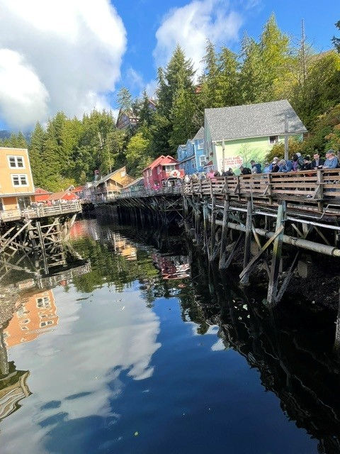 Colorful waterfront boardwalk with people, pastel houses, trees, and blue sky reflected in calm water.