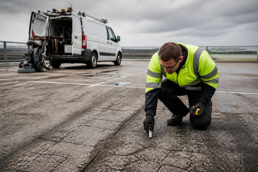 Professional inspector examining car park surface for cracks and wear patterns using measuring tools, with cleaning equipment and maintenance vehicle visible in the background on an overcast day