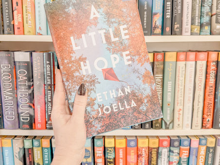 a hand holds a copy of Ethan Joella's A Little Hope in front of a shelf of books