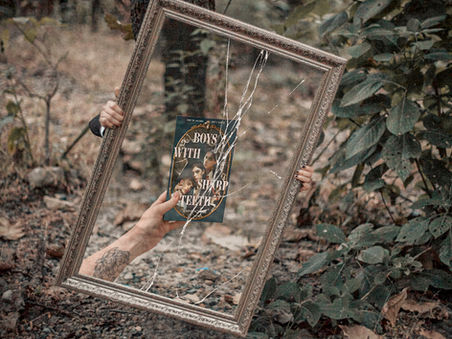 Hands hold a cracked mirror in a forest, reflecting a book titled "Boys with Sharp Teeth." Leaves and earthy tones create a mysterious vibe.