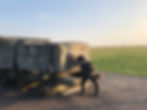 Person looking as if she is pushing large stone block secured with ropes on wooden logs in a grassy field. Sunny, clear sky. Energetic mood.