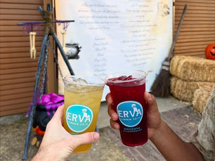 Two hands holding Halloween-themed yerba mate mocktails made with Erva blends, against a spooky backdrop with pumpkins and fall decor.