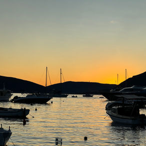 Small boats and yachts anchored in a quiet harbor at sunset, their silhouettes reflected in calm water as golden light fades behind dark mountain hills.