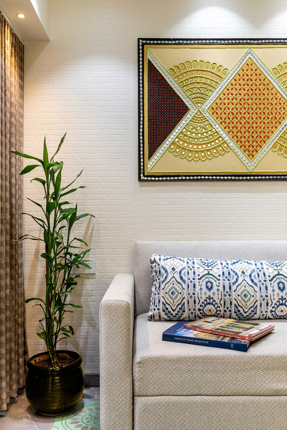 Den in a Hindustani Hygge Mumbai apartment with textured wall art, patterned cushion, indoor plant in a bass pot, and books on a light-toned sofa bed.
