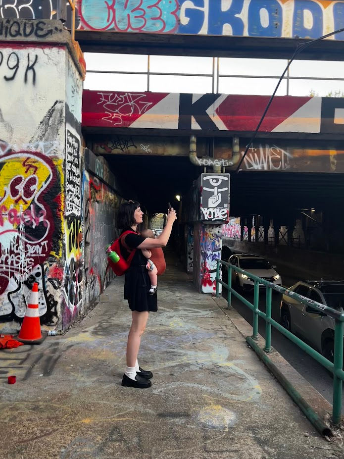 Woman under a graffiti-covered underpass takes a photo with her phone while carrying a baby and red backpack.