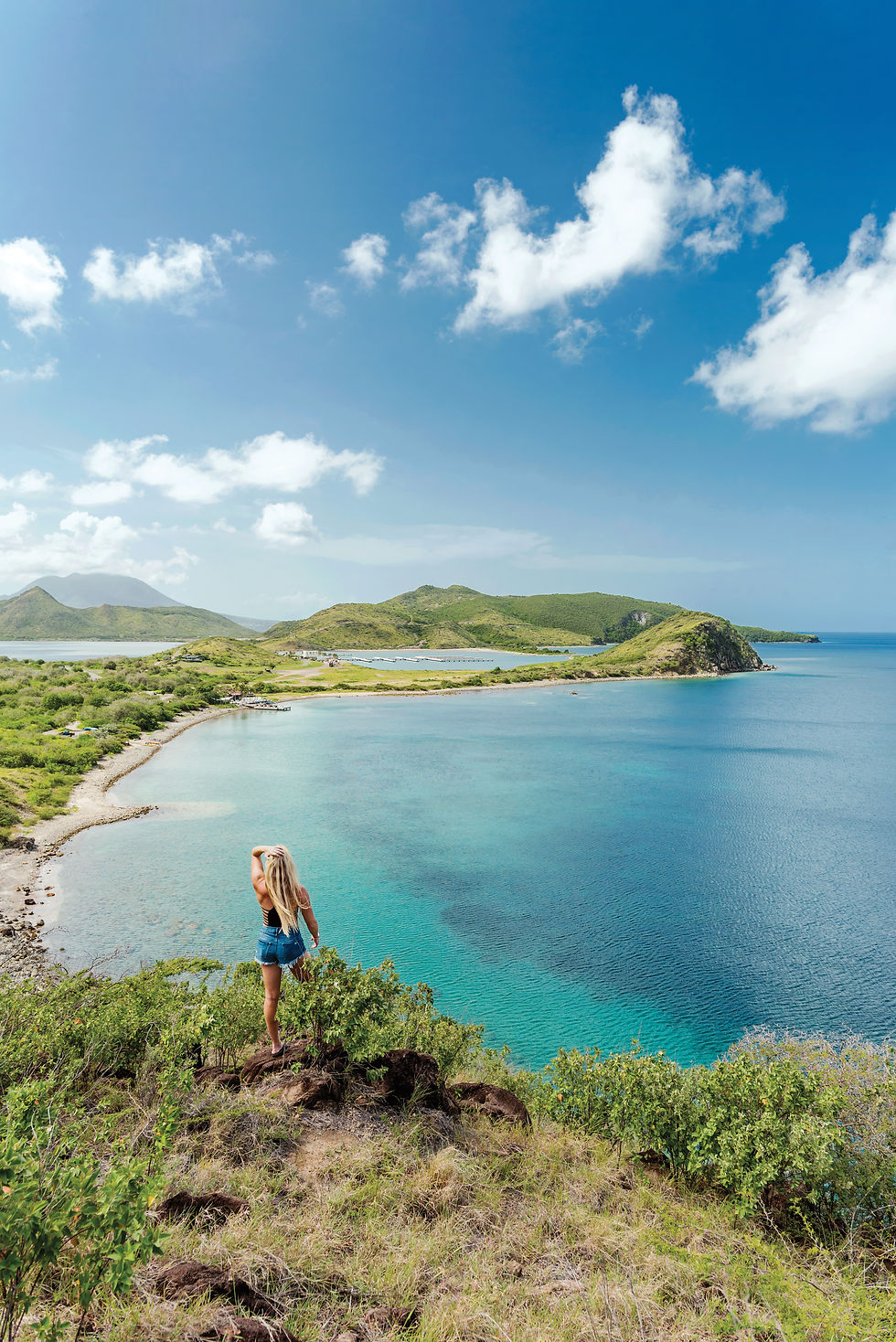 Woman stands on a cliff overlooking a turquoise bay, green islands, and a bright blue sky with scattered clouds.