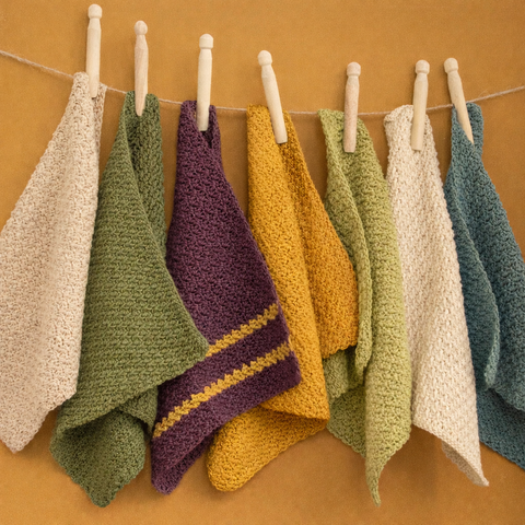 Handgemaakte gehaakte vaatdoeken in natuurlijke Scandinavische kleuren hangend aan een lijn met houten knijpers tegen een warme achtergrond.