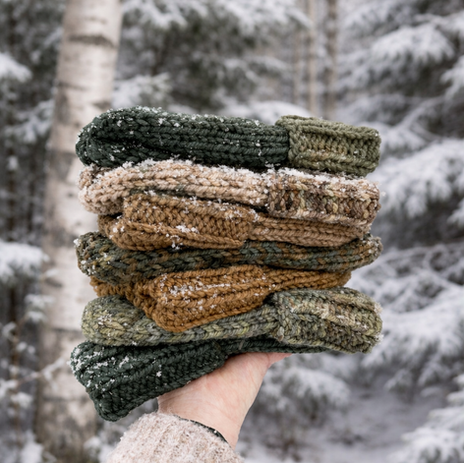 Stapeltje handgebreide wollen mutsen in natuurlijke aardetinten gehouden in een hand met een besneeuwd Scandinavisch bos en berkenbomen op de achtergrond.