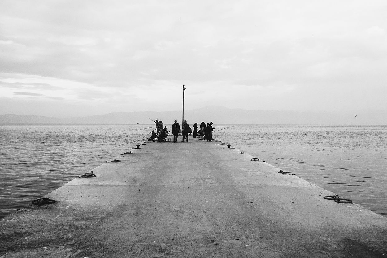 "Barış Gülen siyah beyaz sokak fotoğrafçılığı. Kapalı bir havada denize uzanan uzun beton iskelede balık tutan insanların silüetlerini gösteren atmosferik manzara."