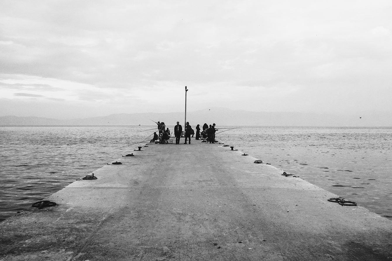"Barış Gülen's black and white street photography. An atmospheric landscape showing the silhouettes of people fishing on a long concrete pier extending into the sea on a cloudy day."