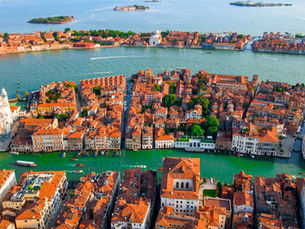 Aerial view of Venice, Italy, showing densely packed terracotta-roofed buildings along bright turquoise canals, with boats moving through the waterways. In the foreground, a large white-domed church stands out among the historic architecture, while across the lagoon small islands and waterfront buildings stretch into the distance under clear blue skies.