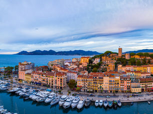 Panoramic sunset view of the Cannes harbor and Old Town (Le Suquet). Rows of white yachts are docked in the turquoise water, framed by warm-toned Mediterranean buildings and a historic hilltop castle under a soft, cloudy sky.