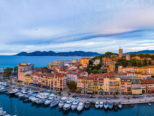 Panoramic sunset view of the Cannes harbor and Old Town (Le Suquet). Rows of white yachts are docked in the turquoise water, framed by warm-toned Mediterranean buildings and a historic hilltop castle under a soft, cloudy sky.