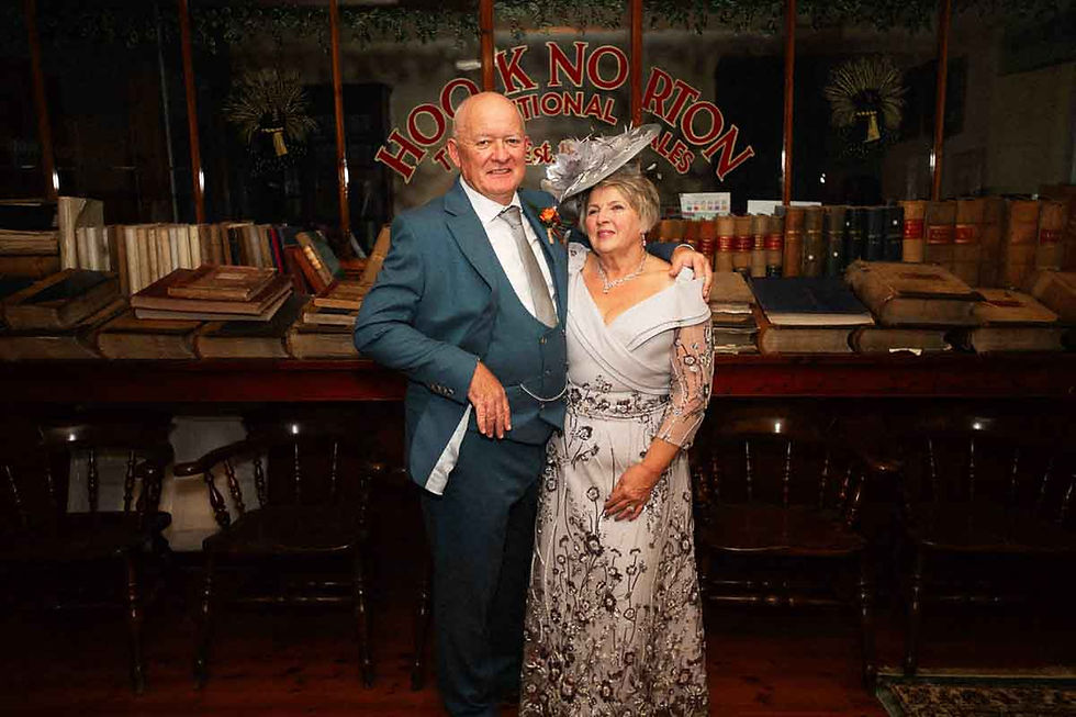 Older couple posing in formal attire in front of books and a Hook Norton sign, smiling at a dimly lit event.
