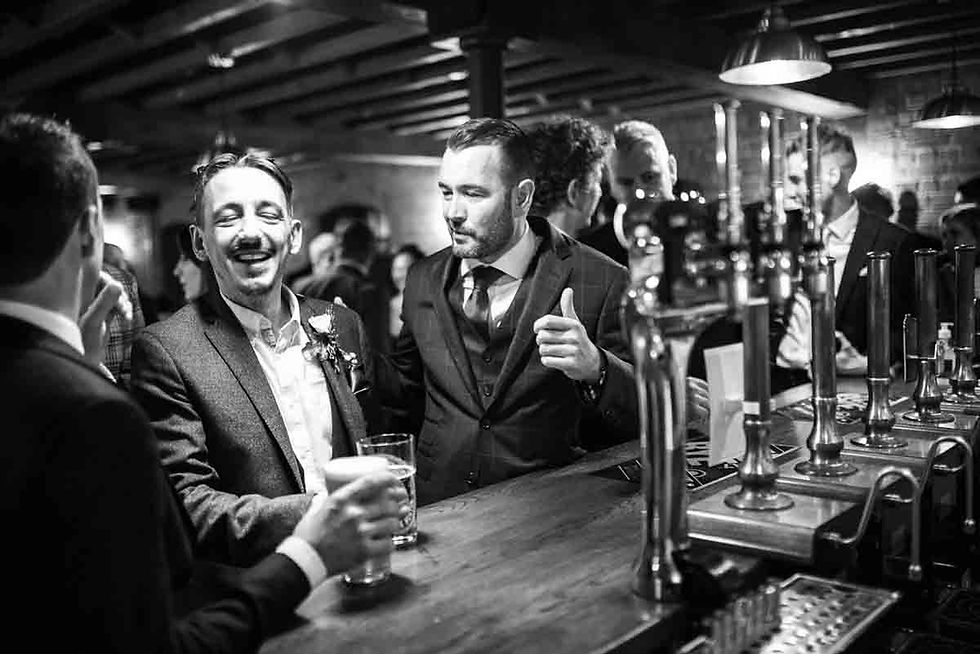 Black-and-white bar scene at Hook Norton Brewery with two suited men laughing and chatting over drinks beside shiny beer taps.