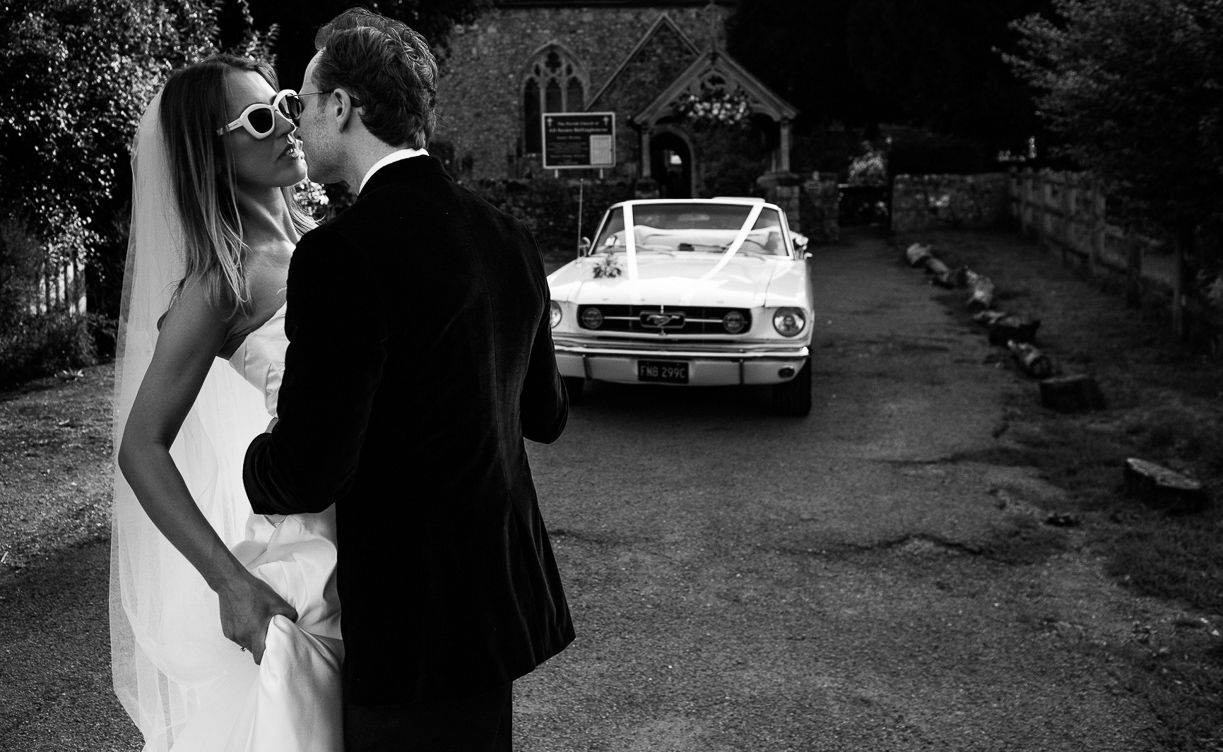 Bride wearing sunglasses about to kiss groom while standing infront of mustang convertible