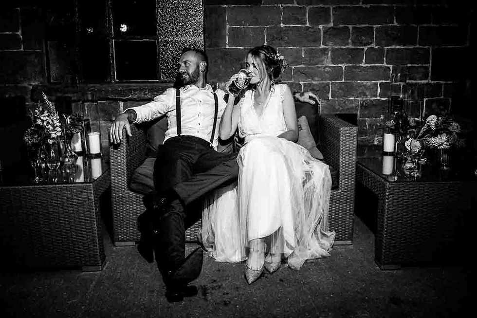 Black-and-white wedding couple lounging on a wicker sofa at Hook Norton Brewery; bride drinks from a glass while groom leans back, smiling.