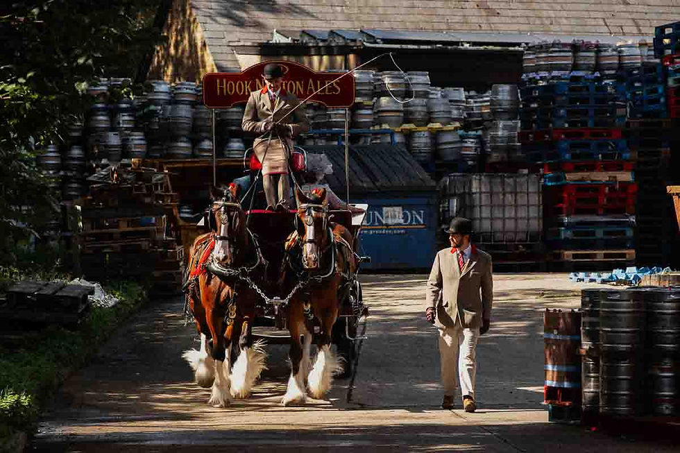 Two shire horses pull a carriage at Hook Norton brewery while two men in suits walk beside stacked beer barrels.