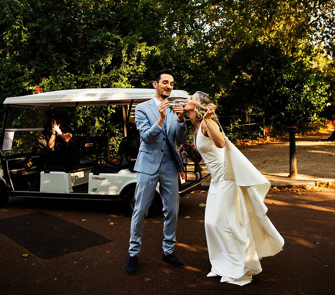 Groom holding champagne glass while bride drinks from it, she is holding her wedding gown train just before stepping into a golf cart to travel to wedding breakfast at battersea park