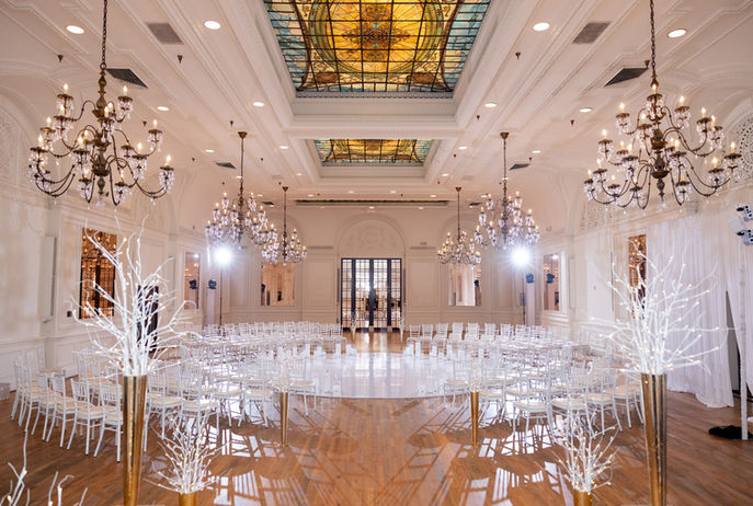 Elegant ballroom with chandeliers, ornate stained glass ceiling, clear chairs setup.