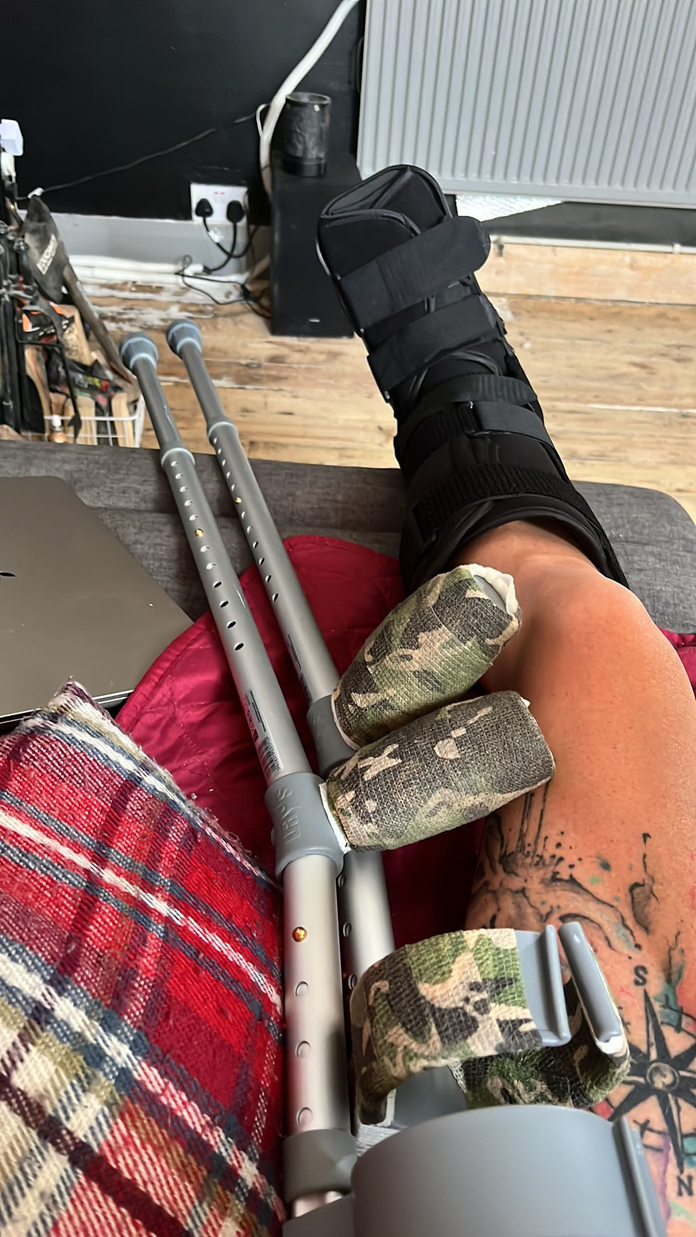 Two crutches with lots of added camo style padding on the handles on a reclining seat with a tattooed leg in a medical boot shows Emma injury.