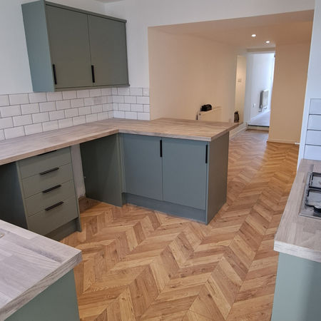 A fitted kitchen with green cabinets, white tiled splashback, and herringbone wood flooring.