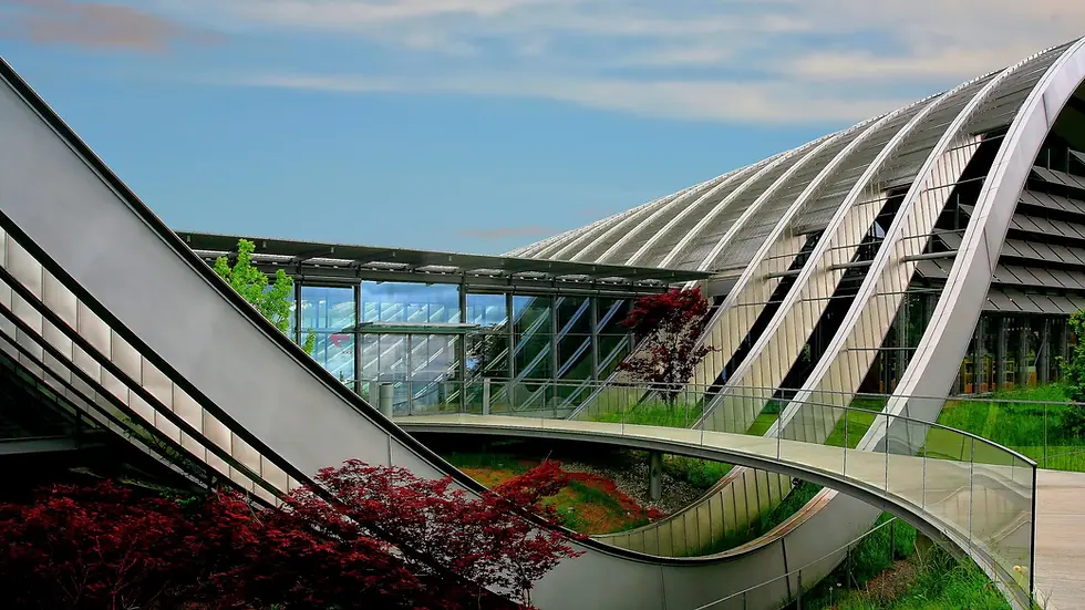 Futuristic glass-and-metal building with sweeping curved roofs and walkways, framed by red shrubs under a blue sky.