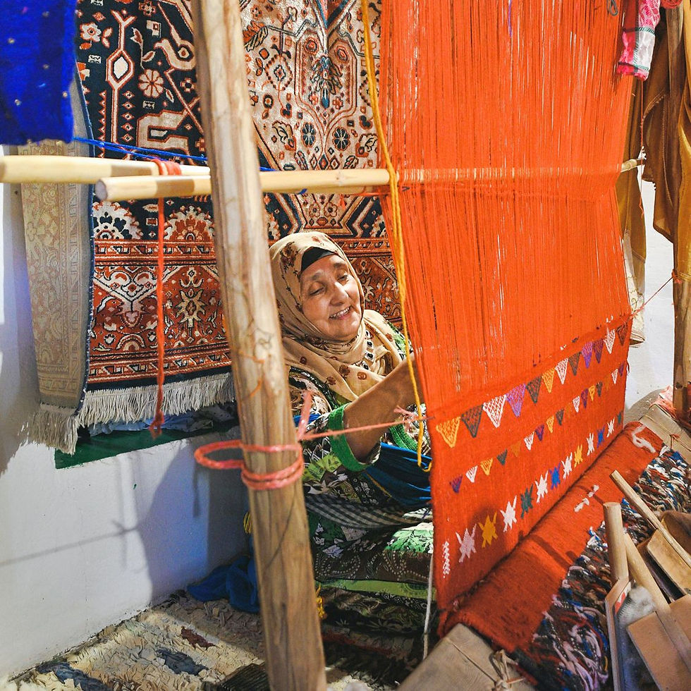 An artisan weaves on a loom with vibrant red and patterned threads. She smiles, surrounded by colorful carpets in a cozy setting.