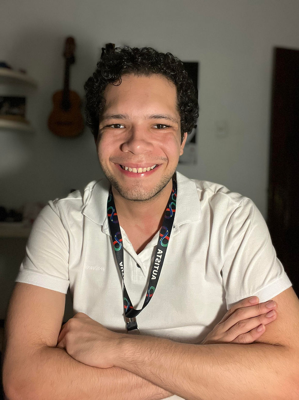 Retrato em plano médio de Lucas Sampaio, o palestrante autista, sorrindo para a câmera com os braços cruzados e vestindo uma camisa polo branca. Ele usa um cordão de identificação com o termo 'AUTISTA' em destaque. Ao fundo, um violão acústico pendurado e prateleiras indicam sua paixão pela música. A imagem promove a neurodiversidade e a visibilidade do autista adulto no Brasil.