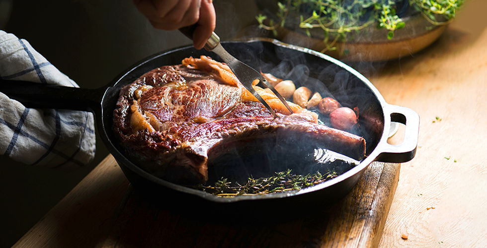 A chef's hand searing a steak professionally in a black skillet with garlic on a wooden table. Steam rises, and herbs are visible. Warm, rustic ambiance.