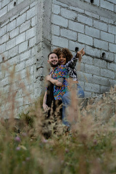 Smiling couple embracing outdoors near a brick wall and tall grass posing together.