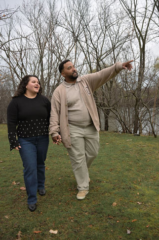 Man points, woman smiles, walking in a park near water and trees.