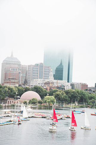 Sailboats on water with a city skyline in the background, cloudy weather.