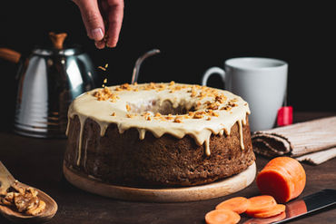 Hand sprinkles nuts on cake, carrot slices, mug, and teapot background.