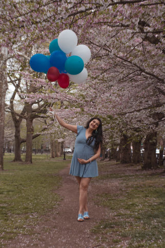 Pregnant woman holding balloons smiling under cherry blossom trees in the park