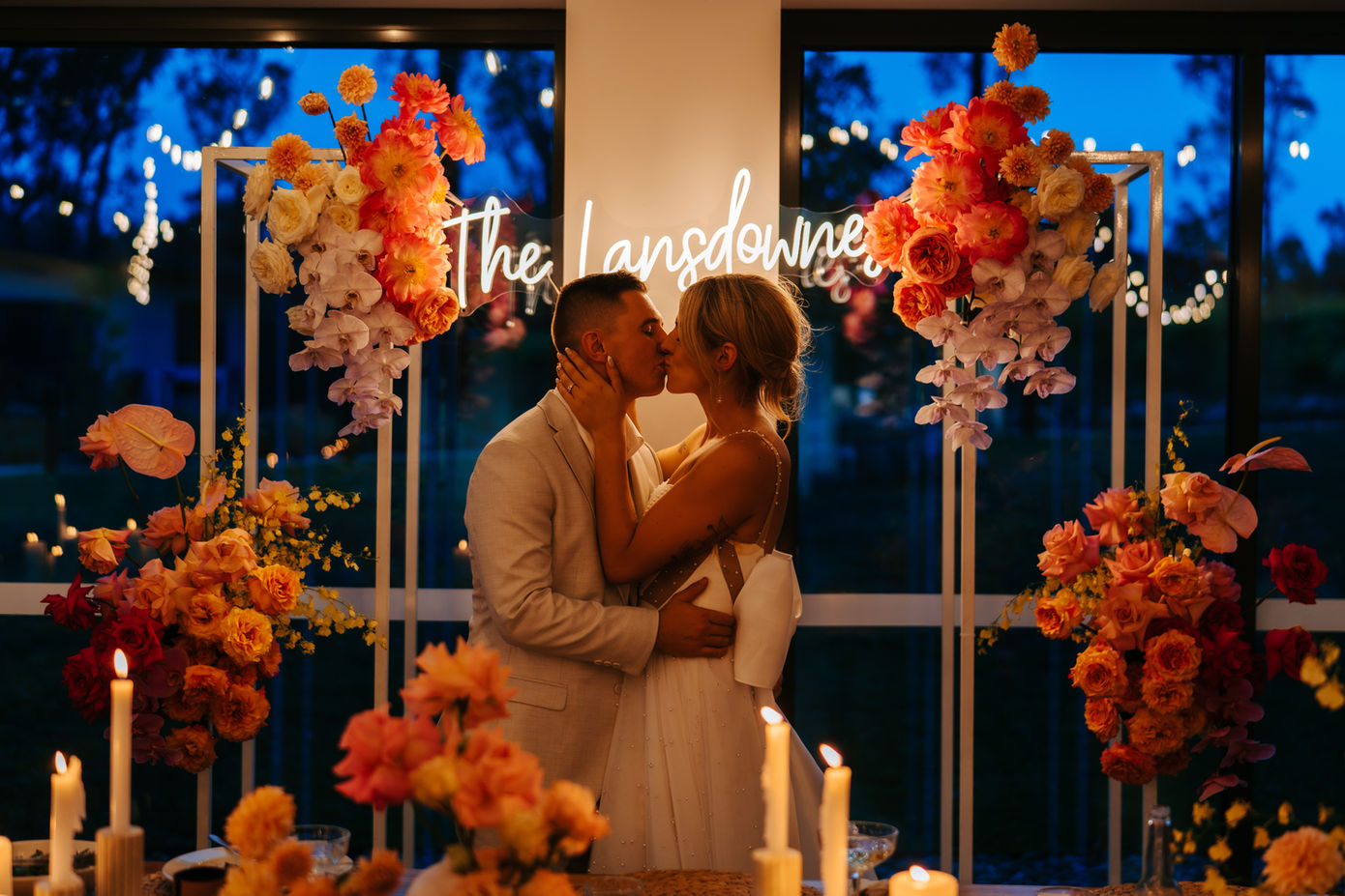 Bride and groom kissing in front of neon sign and flowers at reception.